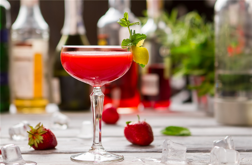 A red cocktail in a coupe glass with ice, strawberries and bottles in the background.
